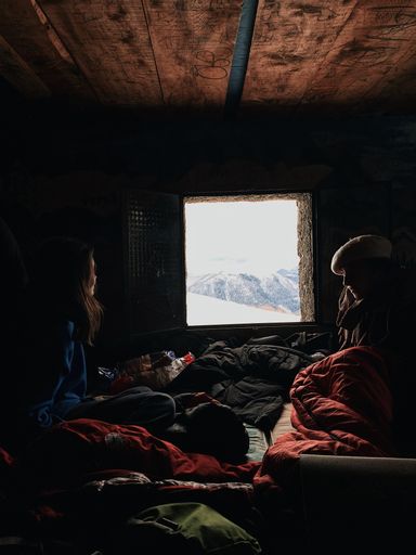 A vertical shot of a pair of female adventurers in a dark cabin in a snowy forest with light shining through an open window