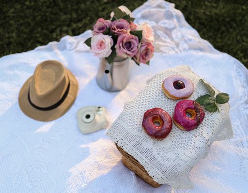 A high angle shot of donuts on a woven basket, a hat and flowers on a white fabric at picnic