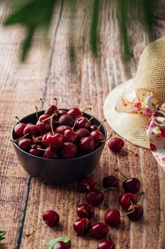 A vertical shot of fresh red cherries on a bowl with a hat and blurred leaves on a table