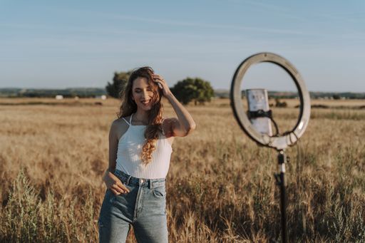 A gorgeous Spanish girl smiling and playing with her hair while taking a selfie on her phone in a field with a ring light in front of her