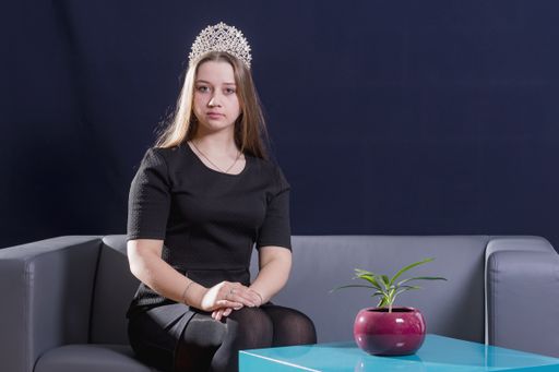 A young beautiful girl in a black dress and a diadem on her head sits on a sofa on a dark background. Hands are folded on the knees.