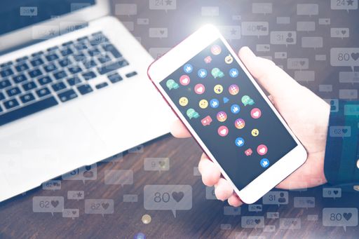 A man’s hand holding a smartphone with reaction emojis on the screen next to an open laptop