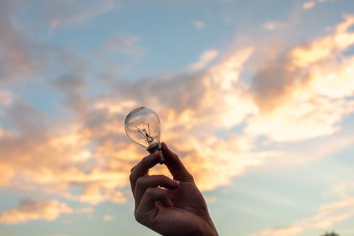 A person's hand holding up a small lightbulb on a beautiful white cloudy sky in the background