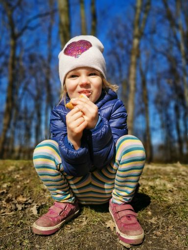 A vertical shot of the girl happily eating her lunch and sitting on the floor in the forest.