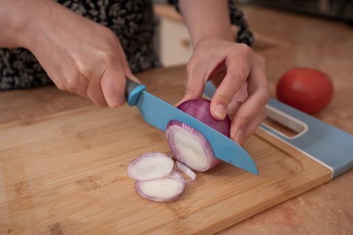 A Close up of woman's hands slicing an onion with a blue knife on a wooden board at the kitchen counter