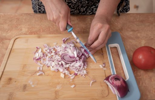 A Top view of a woman's hands chopping onions with a blue knife on a wooden board at the kitchen counter