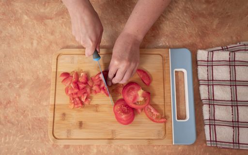 A Top view of woman's hands chopping tomato slices with a blue knife on a wooden board at the kitchen counter