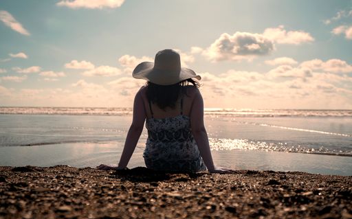 A Hispanic woman sitting alone at the edge of the beach seen from behind, wearing a hat