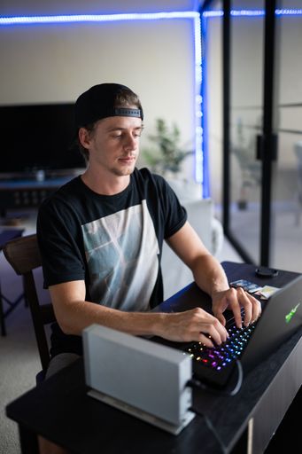 A vertical shot of an American guy with a cap, sitting at his desk and playing on his gaming laptop
