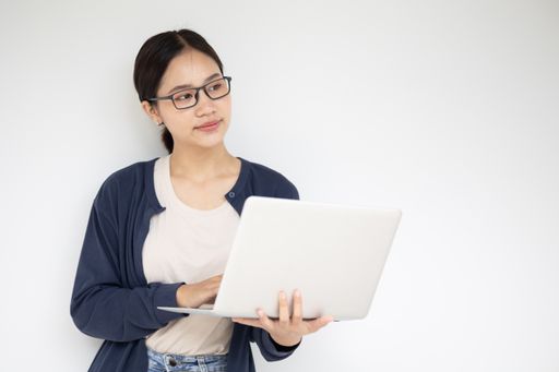 A young Asian girl holding a laptop while thinking standing near a white wall