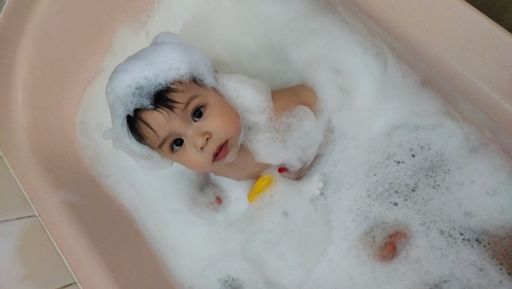 A vertical high angle shot of a cute baby boy covered in soap in the bath, looking up at the camera curiously