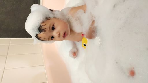 A vertical shot of a cute Swiss-Thai baby boy bathing in a tub, covered in soap with a rubber duck