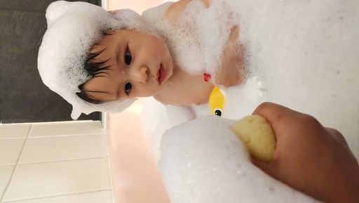 A vertical shot of a curious chubby toddler covered in soap in the bath, looking at a sponge being squished