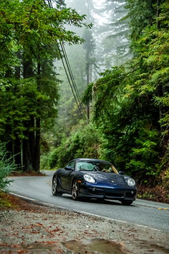 A Dark Blue Porsche Cayman driving on a winding road in the woods on a rainy day