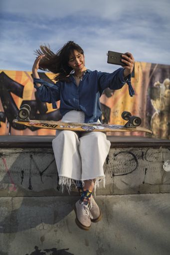 A vertical shot of a girl taking a photo of herself in the park with a skateboard