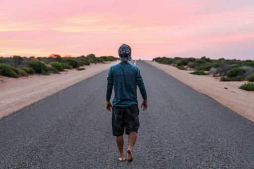 A shot of a barefoot male wearing a cap, blue shirt and shorts walking on a highway road surrounded by greens