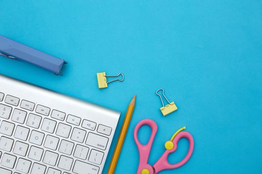 A paper stapler, paper clips, scissors, colored pencil and a keyboard on a blue background