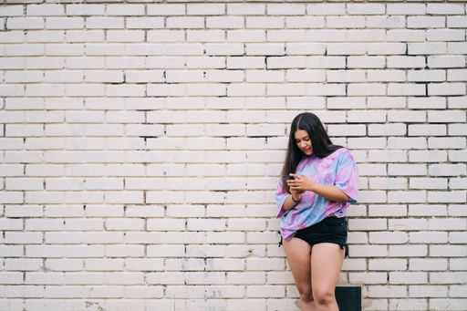 A young chubby Caucasian lady using her phone wearing shorts and a t-shirt leaning on a wall