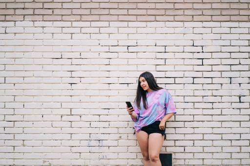 A young chubby Caucasian lady using her phone wearing shorts and a t-shirt leaning on a wall