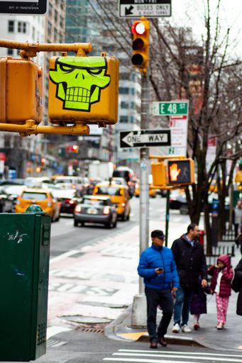 Streets of NYC, man in blue and a father & daughter waiting at a traffic light on foot, cars in the background. Neon skull sticker in the foreground.