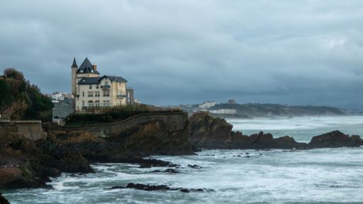 Ocean waves crashing onto sandy beach with houses in the background: Biarritz Palace, France