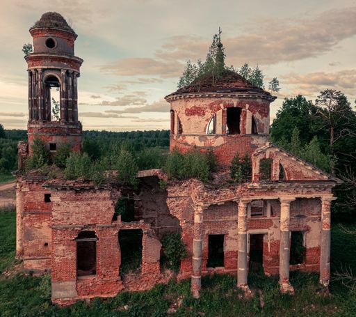 An abandoned church with growing plants on the roof in the forest under a cloudy sky