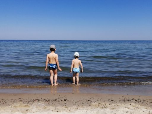 Two young boys standing close by the water on sand at a beach.