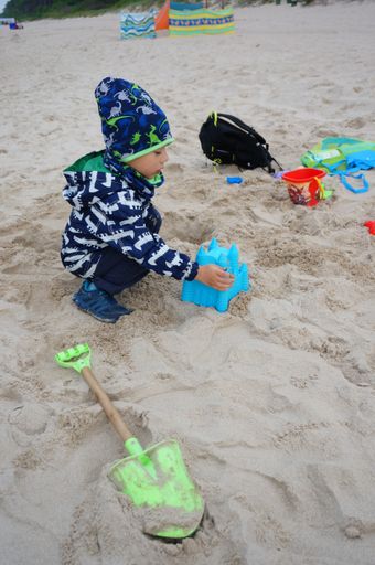 Young small boy playing with plastic toys on sand close by the water at a beach on a cloudy and windy day.