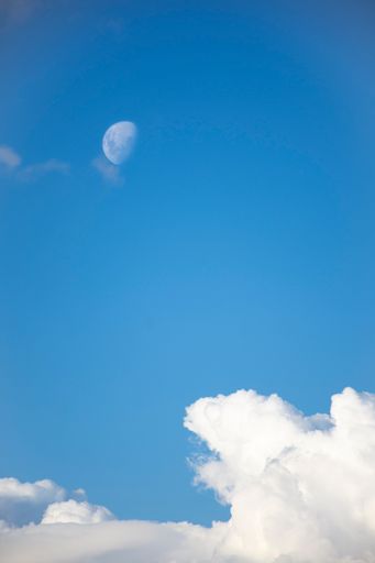 A vertical low angle shot of the moon rising in the cloudy blue sky