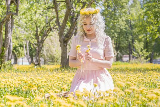A happy young girl in a pink dress and with a wreath of yellow dandelions on her head sits on the lawn and looks at the flowers in her hands.