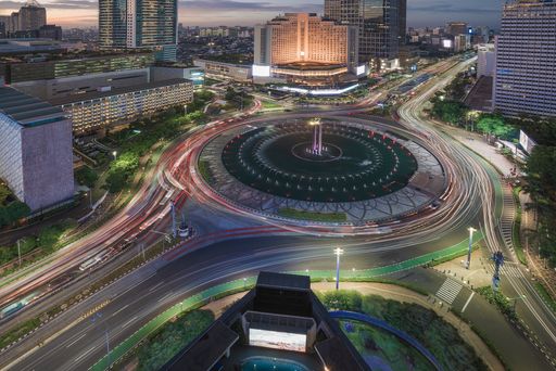 Evening cityscape with a roundabout fountain and car light trails.