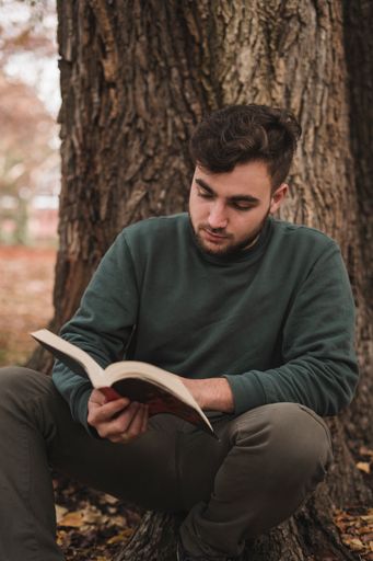 A young Caucasian man sitting next to the tree and reading a book
