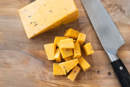 A top view of a knife resting on a wooden cutting board with chunks of English cheddar cheese