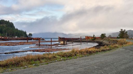 A road beside a log sort leading to the old rusty ship hull breakwater and rain clouds forming in the sky.