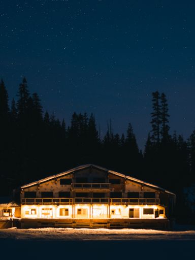 A big house in the woods with lights and silhouette of pine trees in the background under a starry sky