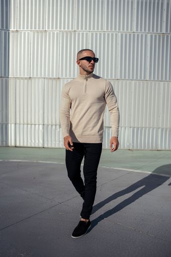 A young man in sunglasses and a beige sweater posing against a white wall on a sunny day.