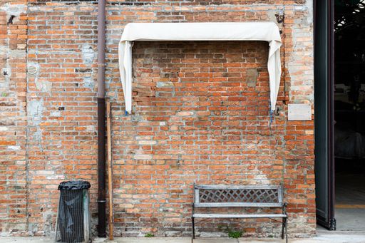 In front of a weathered brick wall, there is an old bench and a garbage collector under a white awning next to an open high gate.