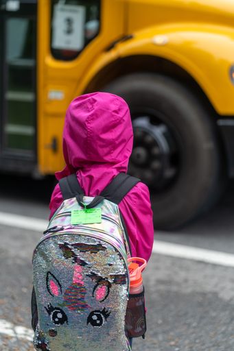 A vertical back view of a little girl with a backpack waiting for the bus