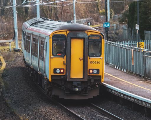 A yellow and silver train arrives at a station with a bridge in the background
