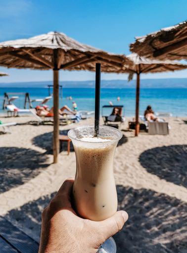 Man holding iced coffee under parasol on sandy beach.