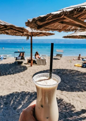 Man holding iced coffee under parasol on sandy beach.