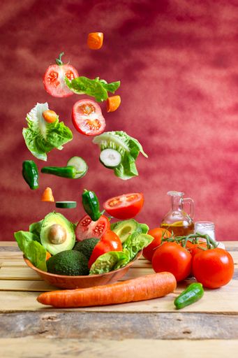 a bunch of vegetables falling on a plate with salad on a wooden table with an oil pan and a salt shaker and bottom