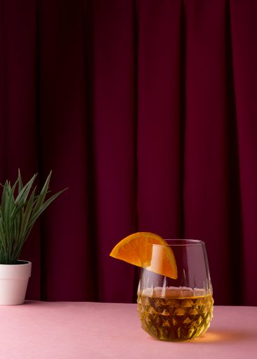 A vertical shot of a glass of drink with an orange slice on a purple curtain background