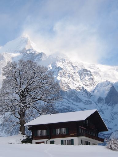 A winter view, with snow, with a wooden house and a huge mountain in the background, in Switzerland