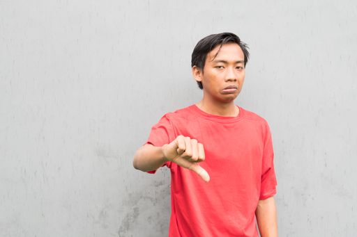An Indonesian guy in a red t-shirt showing thumbs down against a gray wall
