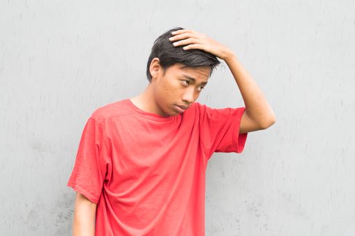 An Indonesian guy in a red t-shirt holding his hair while standing against a gray wall