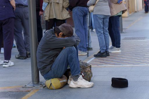 homeless man on the street begging for alms with a sad expression, his head between his legs and a cap on the ground for coins.