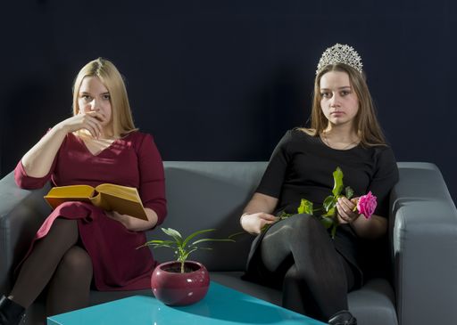 Two girls, blonde and brunette, in red and black dresses, are sitting on the couch. One holds a book in her hands, the other holds a rose