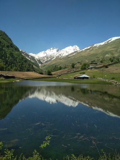 This beautiful photo is from the kinnour valley's sangla kanda lake, India. Surrounded by snowcapped mountains and green tree makes it panoramic view.