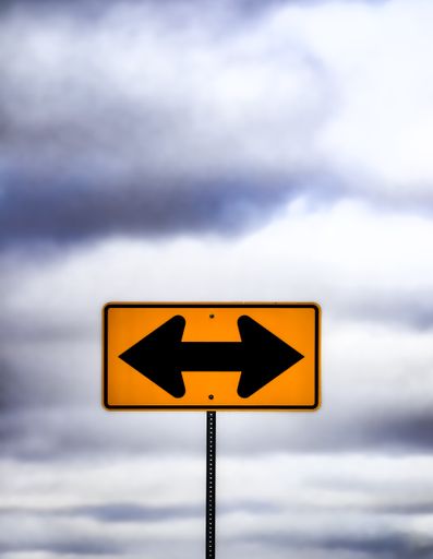 An arrow-shaped street sign against cloudy sky at corner intersection